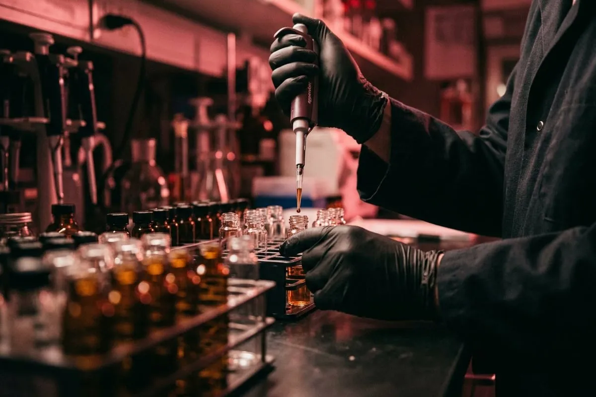 科學家雙手在昏暗實驗室中以移液管操作小玻璃瓶，暖玫瑰色背光投射